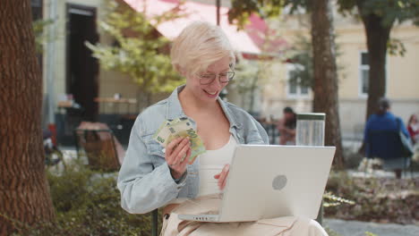 happy rich young woman using laptop winner victory holding cash money in euro banknotes in city