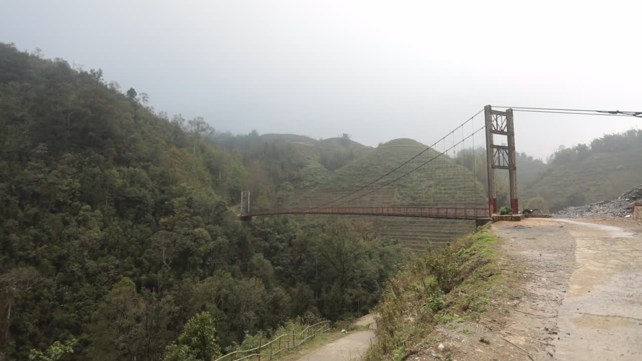 A misty suspension bridge spans a lush, mountainous valley in Sa Pa, Vietnam under cloudy skies