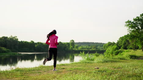 a girl in a pink jacket and black pants runs near the river in headphones preparing for the marathon