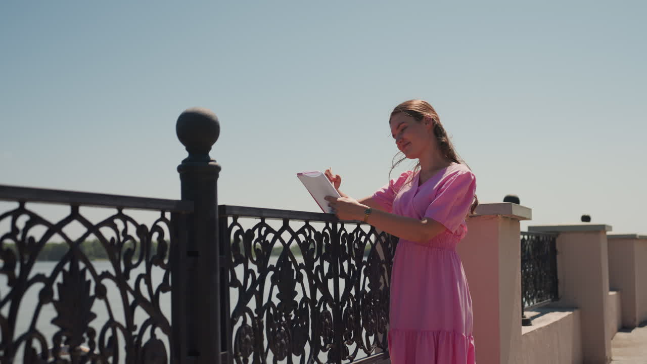 Mulher de vestido rosa na sacada lendo uma carta sob um céu limpo, grade de ferro ornamentada, terraço à beira-mar iluminado pelo sol, brisa suave, clima contemplativo, envelope vintage, enquadramento cinematográfico, atmosfera romântica