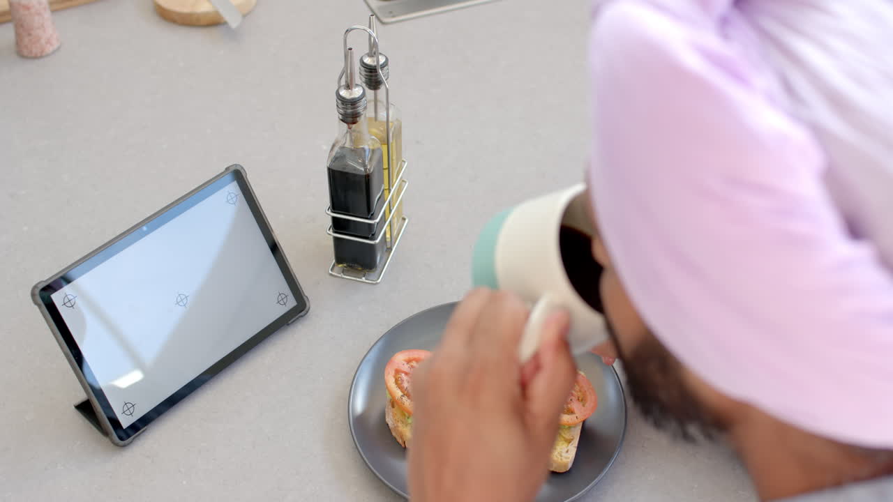 Drinking coffee, man in turban watching tablet while eating breakfast at home kitchen
