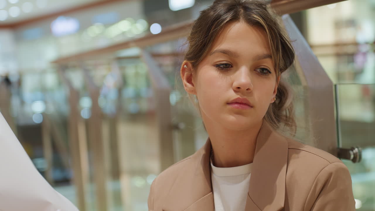 Individuo reflexivo bajo luces suaves, figura contemplativa sentada junto a una barrera de cristal, creativo caucásico que parece introspectivo mientras se relaja en un centro comercial suavemente iluminado