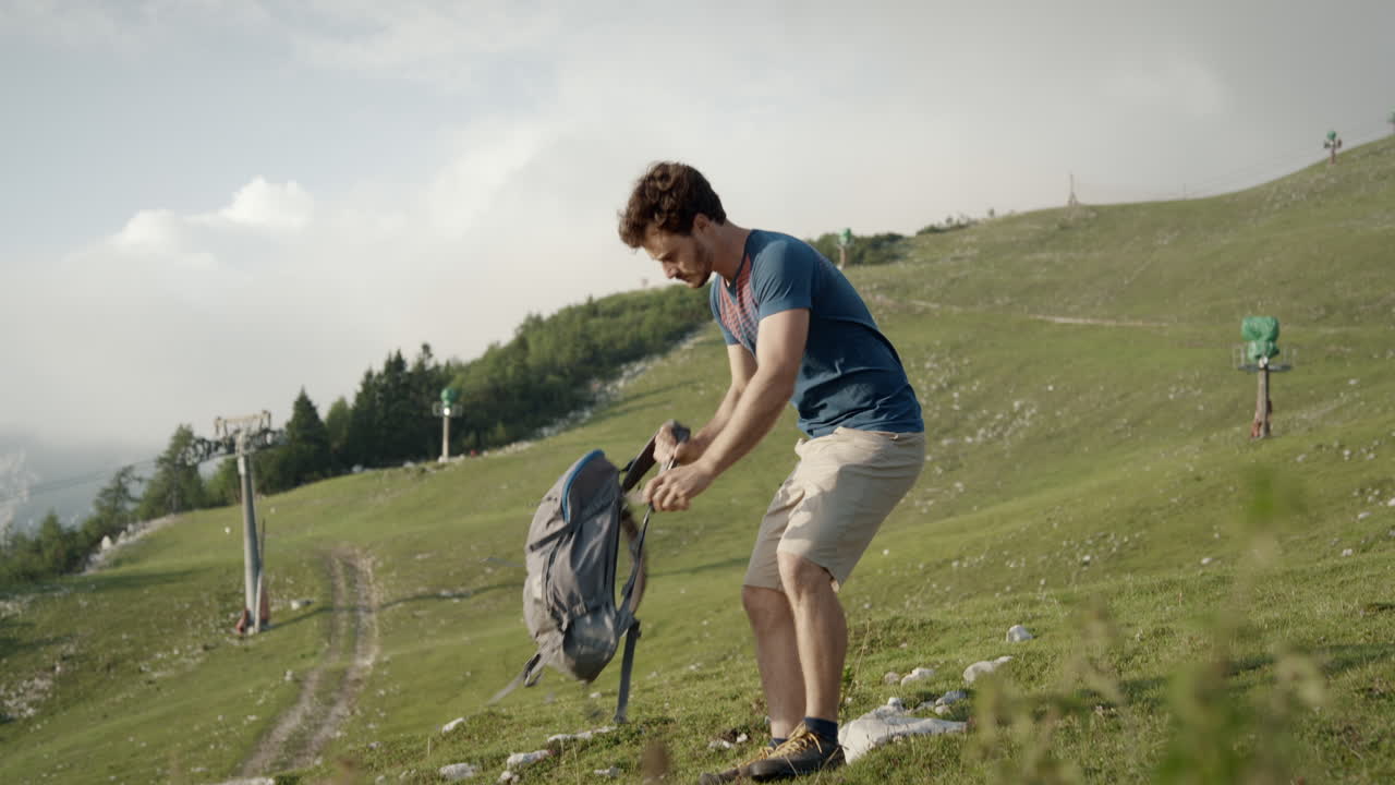 un joven excursionista sube una colina, se detiene, se sienta y abre su mochila para sacar un pequeño folleto