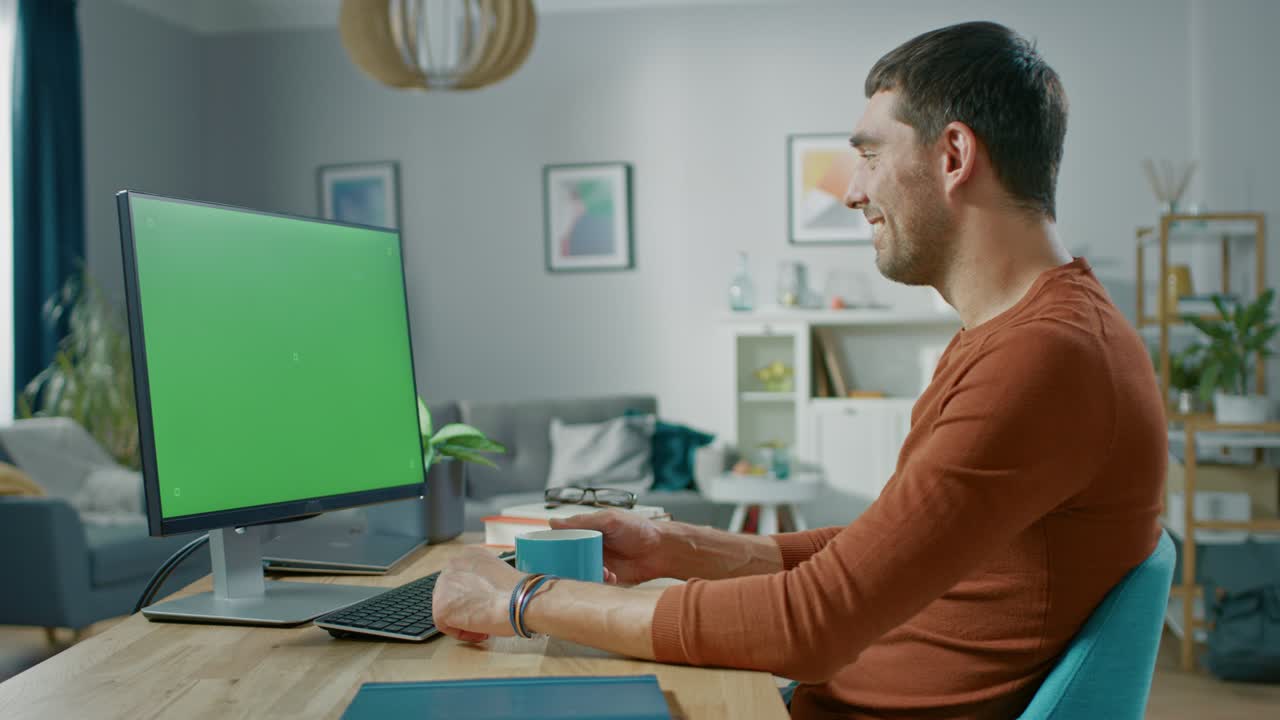 un bel uomo sorridente seduto alla sua scrivania a casa usa un personal computer con un modello di schermo verde. vive in un accogliente appartamento con un moderno interno in salotto.