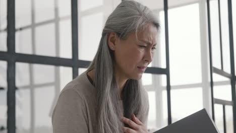 Middle-Aged-Woman-Having-Chest-Pain-While-Reading-A-Book-Sitting-On-Sofa-And-Then-Standing-Up