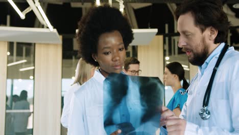 Caucasian-and-african-american-doctors-in-medical-masks-speaking-and-discussing-Xray-scan-in-hospital-office