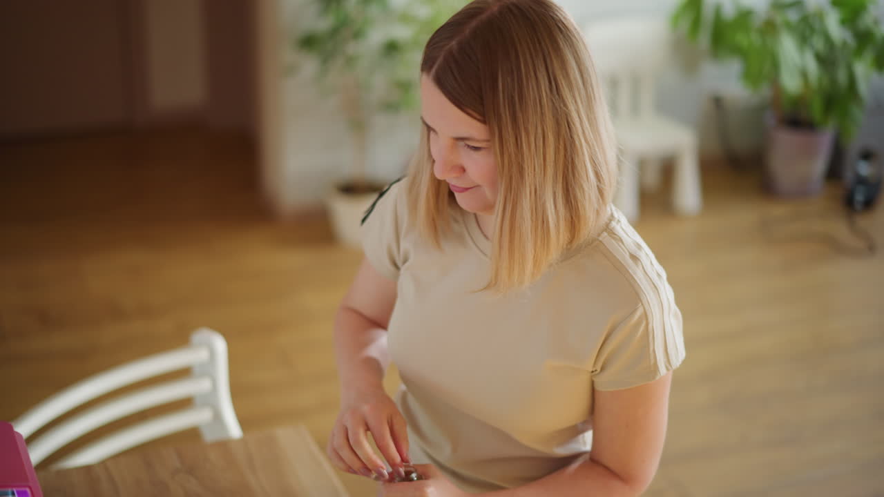 Smiling woman standing in bright home interior holding small object in hand, wearing casual striped pants and beige t-shirt, wooden floor, potted plants and robotic vacuum visible