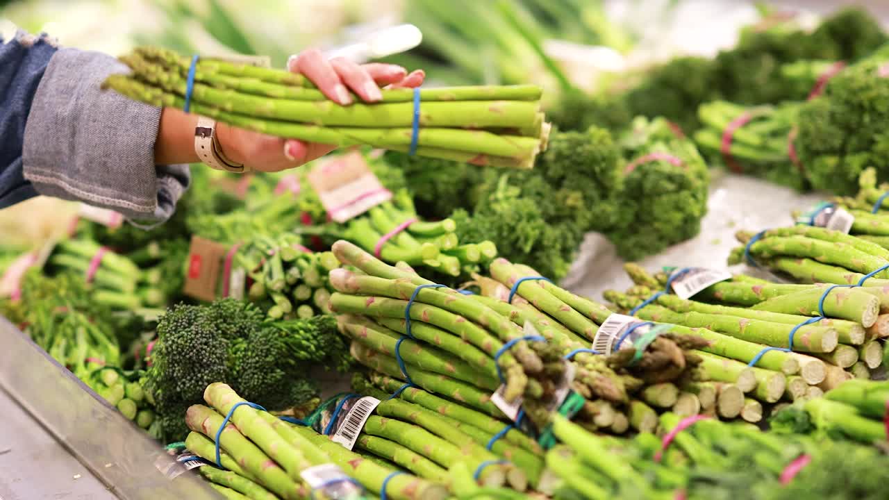 A person examines asparagus bundles in a well-lit supermarket produce section, focusing on freshness and quality