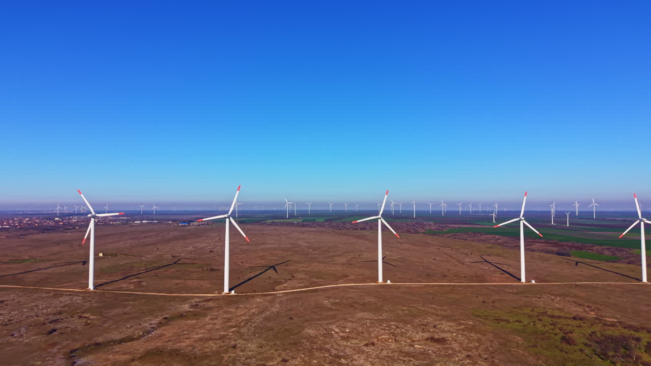 As turbinas eólicas geram energia em um dia claro, em um grande campo aberto com um céu azul e colinas distantes no plano de fundo.