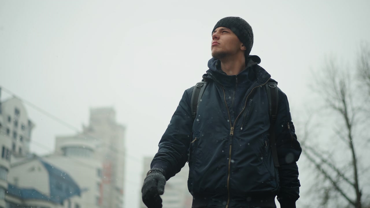Man wearing dark winter clothing and backpack walking outdoors in snowy urban environment with tall buildings and bare trees visible. Snowflakes fall around him creating cold atmosphere
