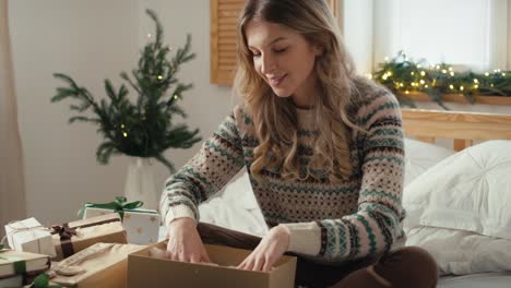 mujer caucásica sentada en la cama y empacando el regalo de navidad en el dormitorio.