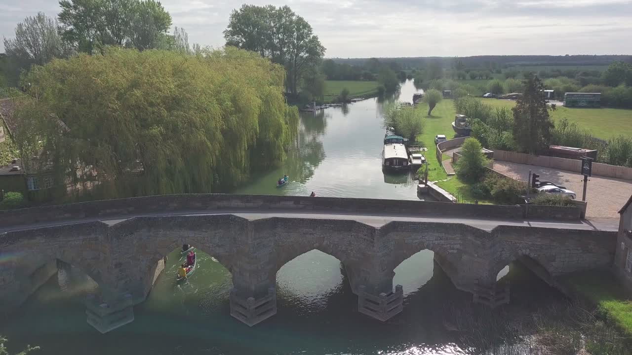 Group Of People Paddling Kayak And Passing Under The Stone Bridge In River Thames With Green Nature Scenery In Abingdon Town Near Oxford City, UK. - aerial drone shot