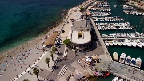 Toma-Inclinada-De-Elevación-De-Drones-De-Un-Día-Ajetreado-En-La-Playa-Junto-Al-Puerto-Deportivo-En-Cassis,-Francia