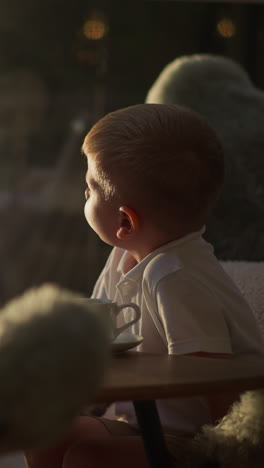 children sit at table in tent. siblings drink tea looking out of sunlit window in hotel slow motion. calm kids rest in glamping tent in morning