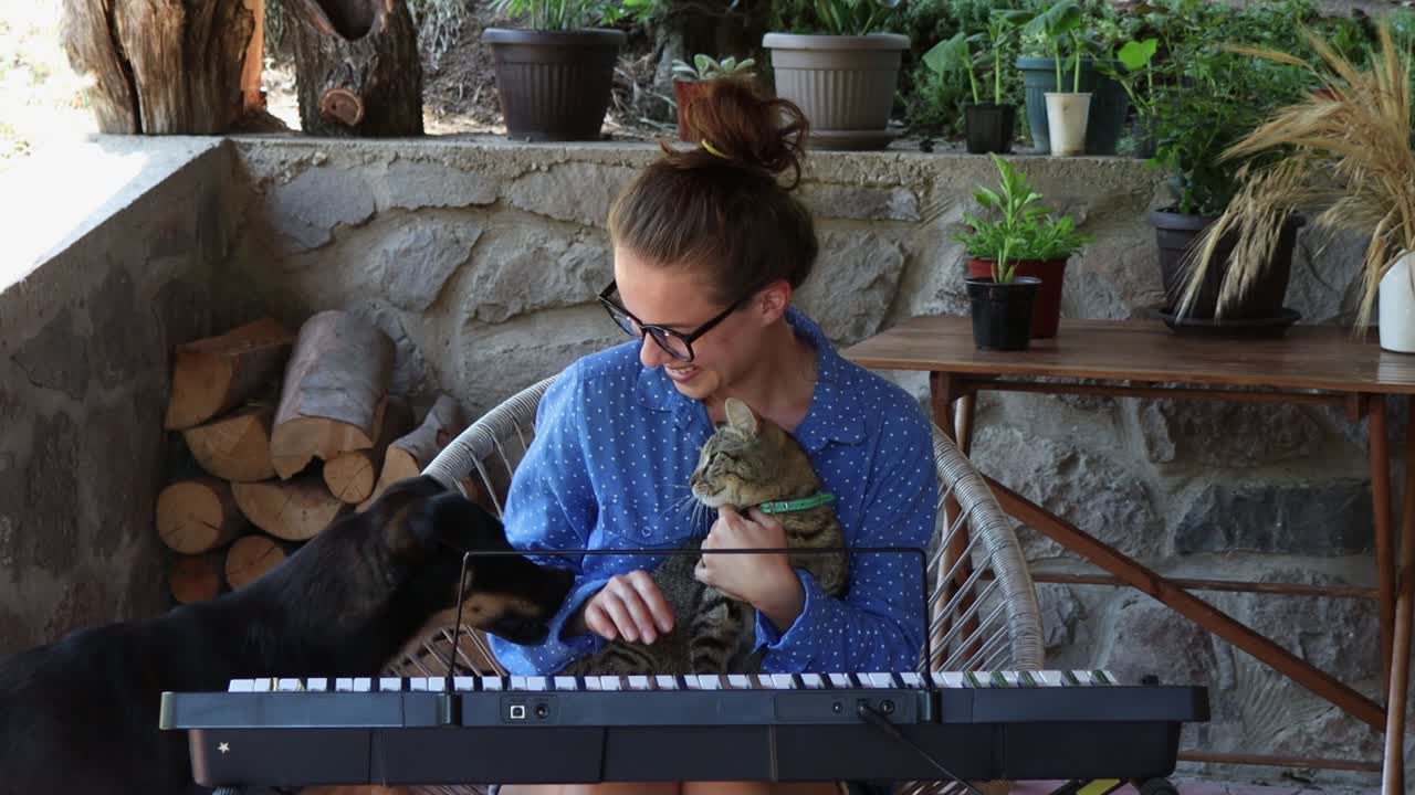 una mujer joven con gafas se divierte con su gato y su perro mientras juega al sintetizador en el patio en el campo.