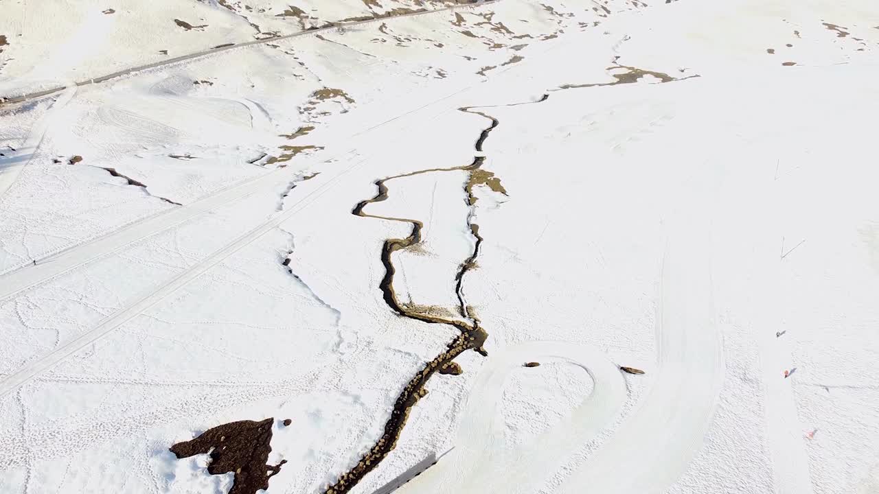 Drone shot of a Stream in the middle of a Snowy Mountain Plain with a Road Behind
