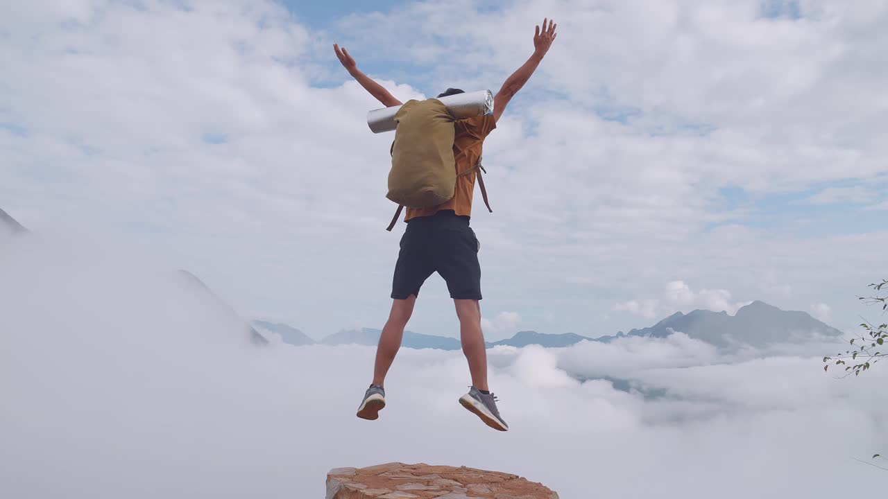 Escursionista asiatico in piedi sulla roccia che salta felice e alza le mani celebrando il raggiungimento della cima di una montagna nebbiosa.
