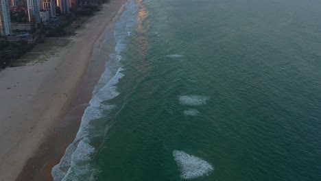 Cinematic-birds-eye-view-of-beautiful-long-stretch-of-sandy-beach-at-Broadbeach,-Gold-Coast,-aerial-sunset-view-of-waves-crashing-the-shore-with-waterfront-high-rise-buildings-along-the-coastline