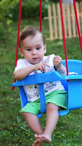 lovely boy in white t-shirt and green shorts outdoors. little baby swaying in the plastic swing tied to the tree. garden backdrop. vertical video