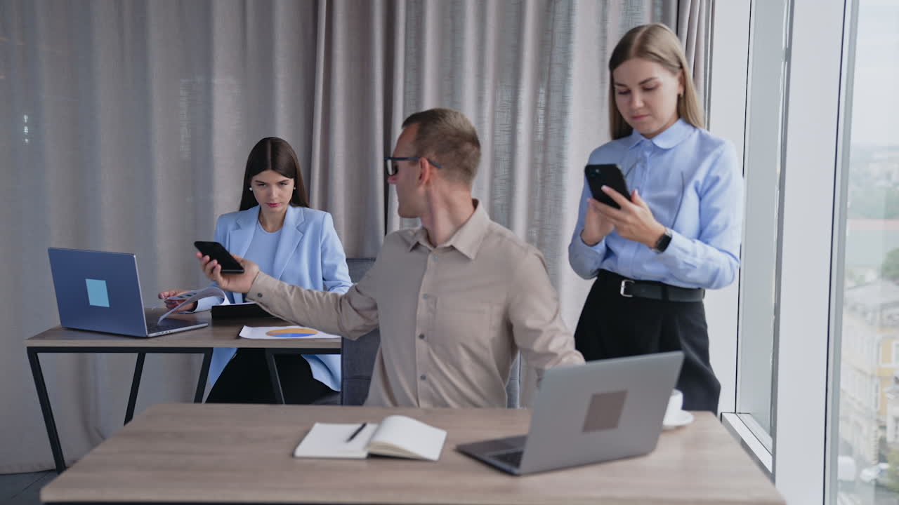 Team of colleagues in the light office room. Man sitting at computer takes phone and passes it to female teammate. Blonde lady with phone in hands standing beside male employee.