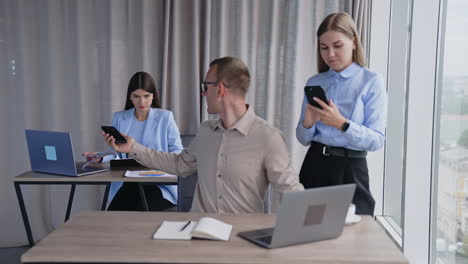 team of colleagues in the light office room. man sitting at computer takes phone and passes it to female teammate. blonde lady with phone in hands standing beside male employee.