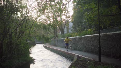 man in yellow jacket runs alongside river with playful dog.