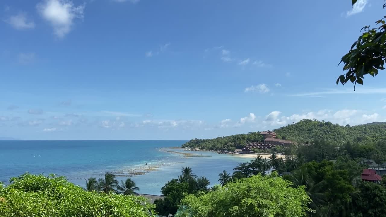 Sweeping view of tropical bay in Koh Samui, Thailand with turquoise water, palm-covered hills, and resort nestled in lush greenery under bright sky from elevated angle