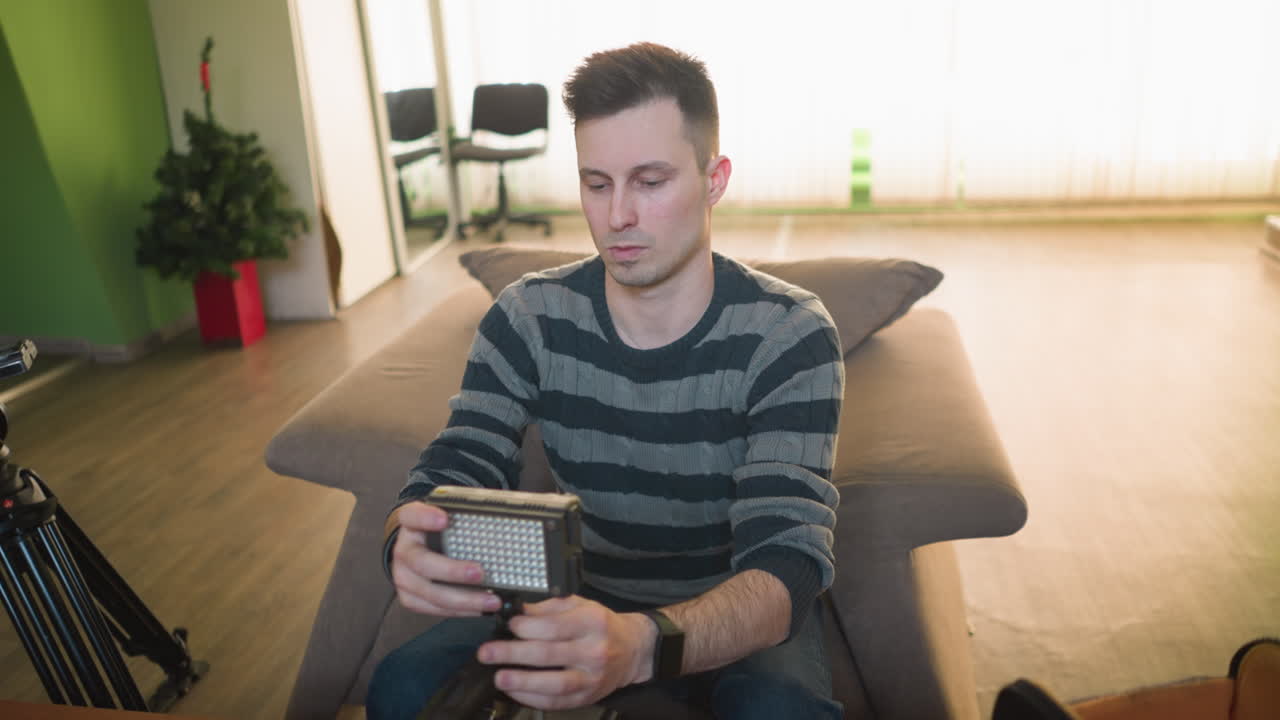 Person holding LED light panel while sitting on sofa in home studio, checking lighting equipment. Preparing for filming, surrounded by camera gear, microphone, and other equipment