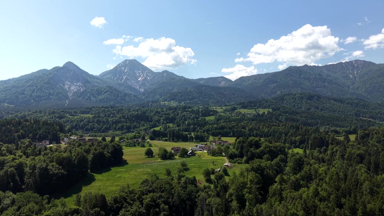 Alpejski las pokrywa dno doliny, podczas gdy wijąca się rzeka, tor kolejowy i linie energetyczne rozciągają się w kierunku odległych, ośnieżonych Alp Austriackich pod letnim niebem. Austria. Ujęcie wprowadzające z drona.