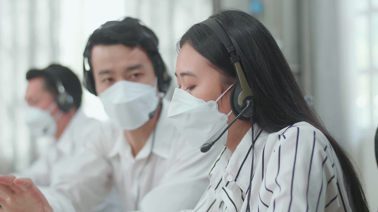 Close Up Of A Man And A Woman Of Three Asian Call Center Agents In Headsets And Masks Discussing About Work While Their Colleague Is Speaking With Customer On The Call At The Office