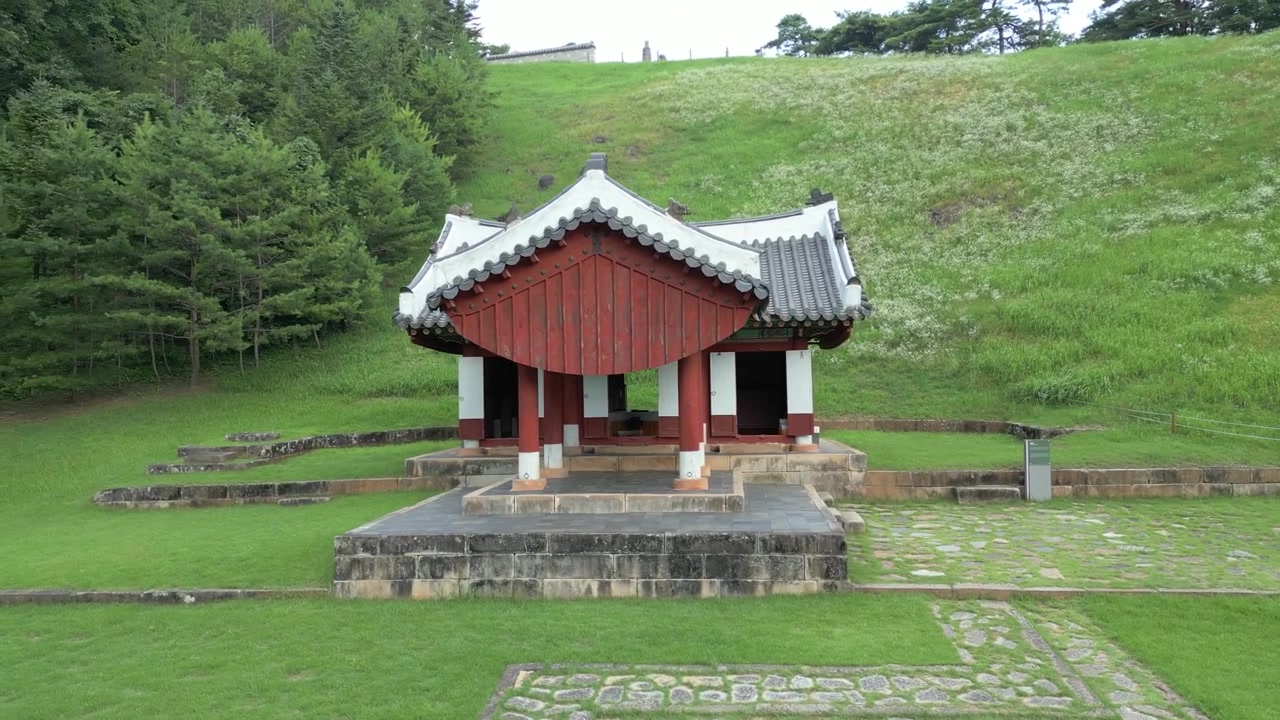 Filmisk udsigt fra jordhøjde af en traditionel koreansk pavillon omgivet af velplejede græsplæner og skovklædte bakker inden for et historisk kulturarvssted