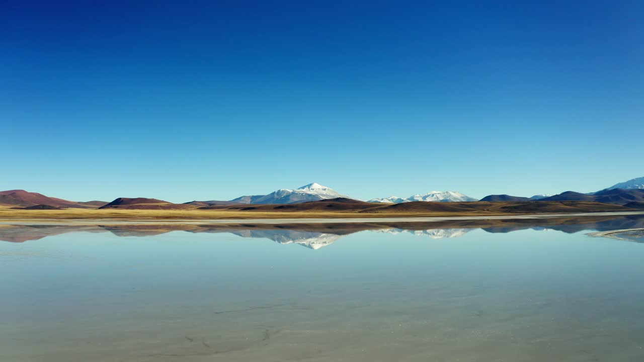 Low aerial tilt up across calm shallow waters and salt crusts at Laguna Brava reflecting snowy mountain, La Rioja Argentina