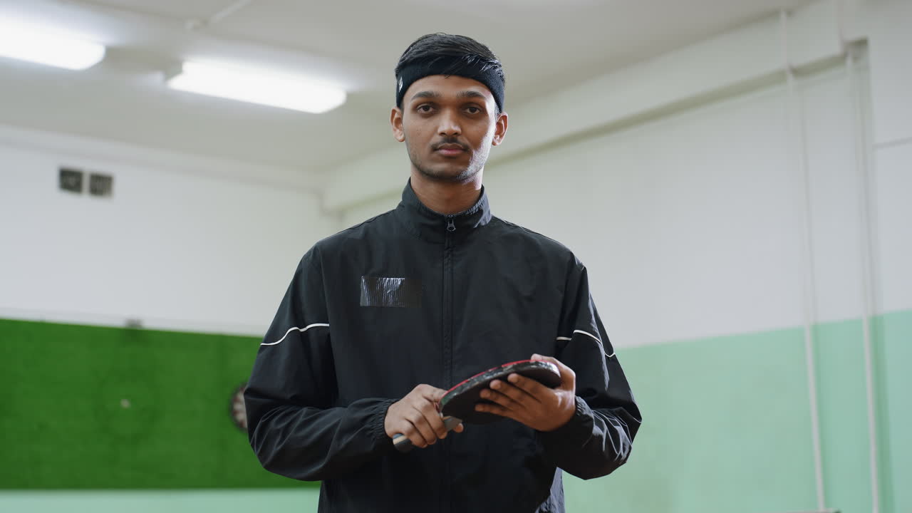 Calm tennis player in black tracksuit holds racket confidently and flips it on palm during indoor practice session, maintaining composure, and concentration in sports hall with green background