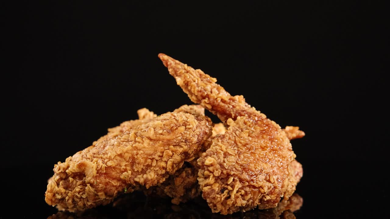 Three crispy fried chicken wings rotate smoothly on a glossy black surface with dramatic studio lighting, highlighting texture and golden color in a seamless loop