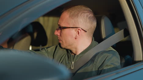 a close-up front view of a man wearing glasses and a green jacket, seated in the driver s seat of a car. he appears focused, looking straight ahead, with a serious expression