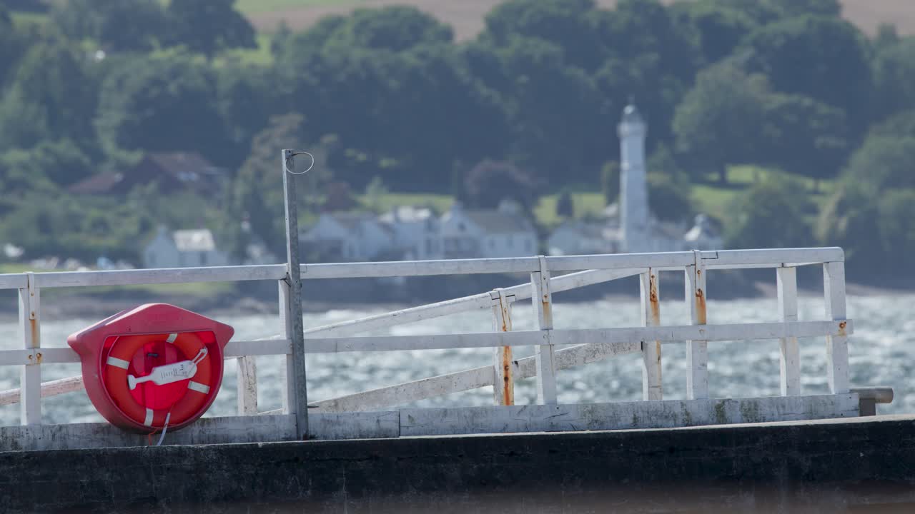 Daylight pan reveals lifebuoy, pier fence, river, distant lighthouse, village, and green hills