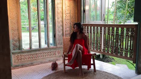 young asian girl in long red dress sitting in a traditional bohemian villa looking out the window towards the garden in bali