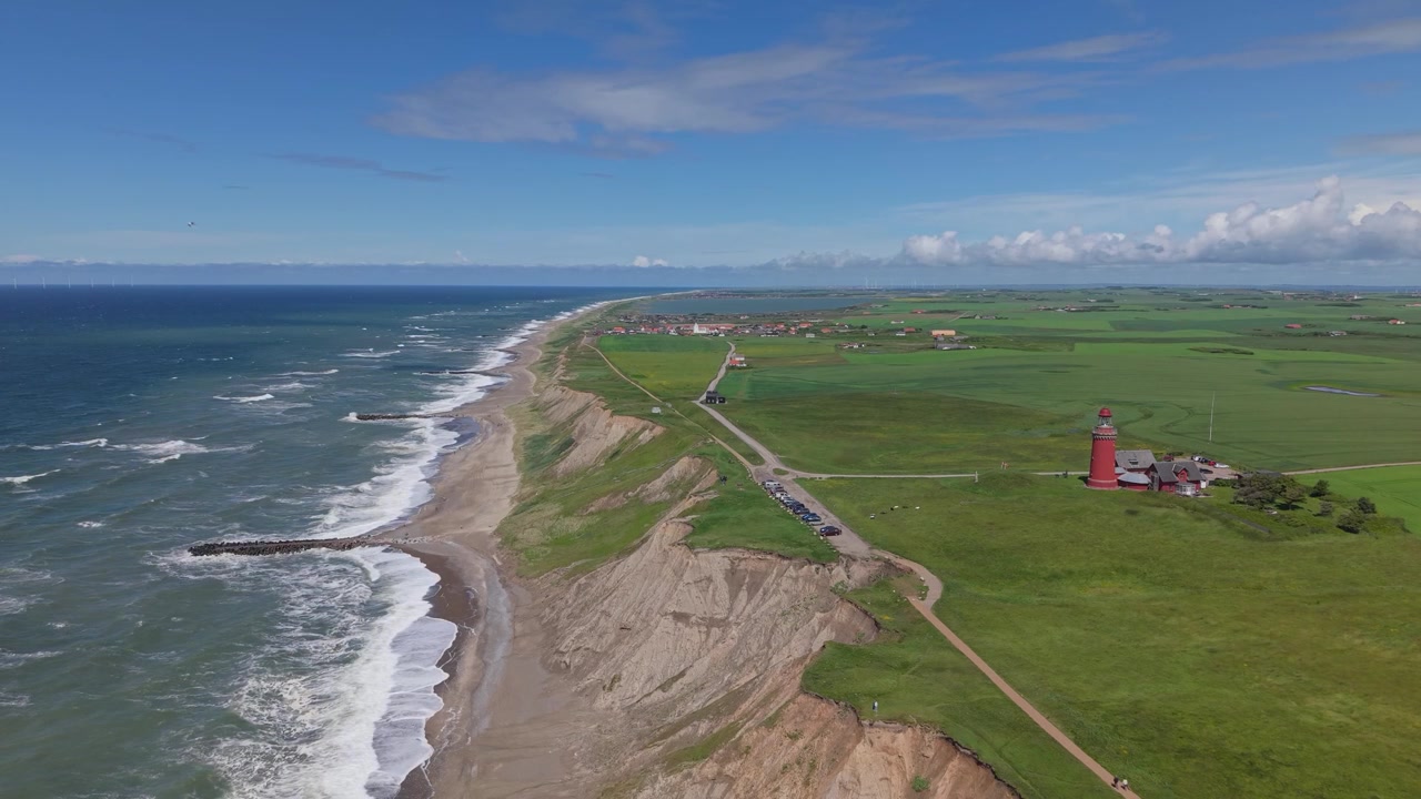Statisk panoramisk flygfilm som fångar den vilda västra Jyllandskusten en blåsig dag, med rullande Nordsjövågor, gröna ängar, Bovbjerg fyr och Ferring by synliga vid horisonten.