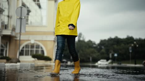 A-sad-teenage-girl-in-a-yellow-jacket-and-orange-rubber-boots-walks-better-cutting-through-the-water-and-is-sad-on-the-street-after-the-rain