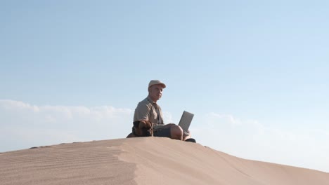 male traveler sitting on sandy dune with dog and using laptop during vacation in sunny day