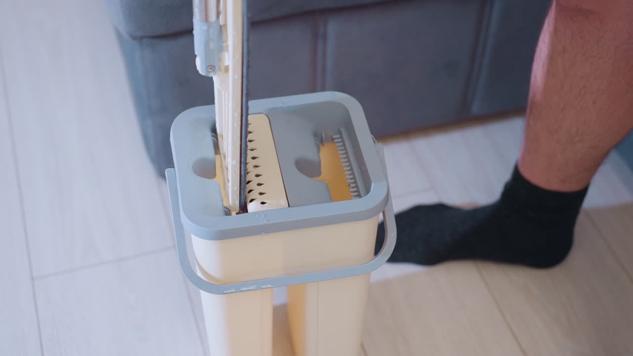 Close up of mop handle pressed into cleaning bucket with built-in wringer while person wearing black socks stands on tiled floor, focusing on detailed view of household cleaning process