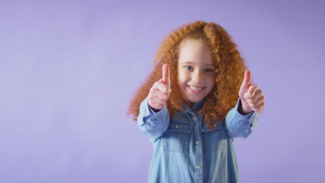 foto de estudio de una chica sonriente con el cabello rojo dando pulgares dobles contra un fondo púrpura