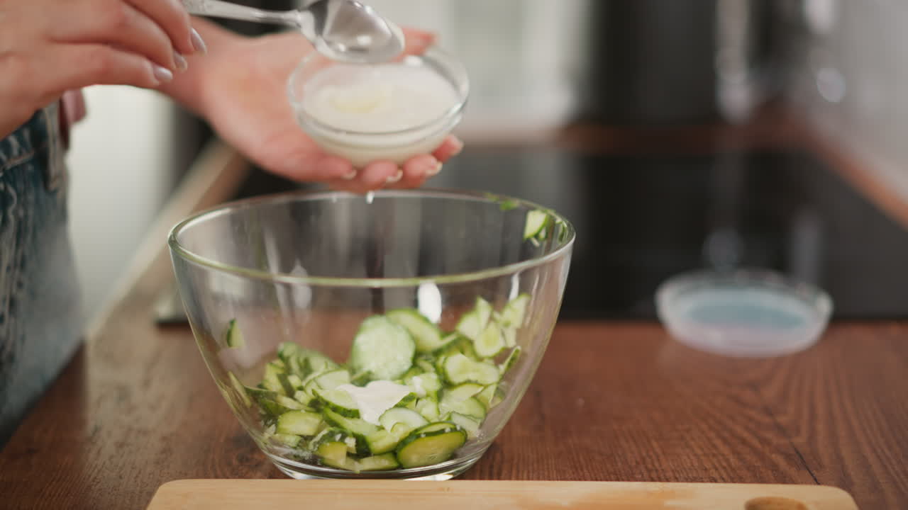 close up of person holding spoon of tangy white sauce over glass bowl of sliced cucumber; drizzle sauce onto crisp green slices while knife and cutting board blur in background