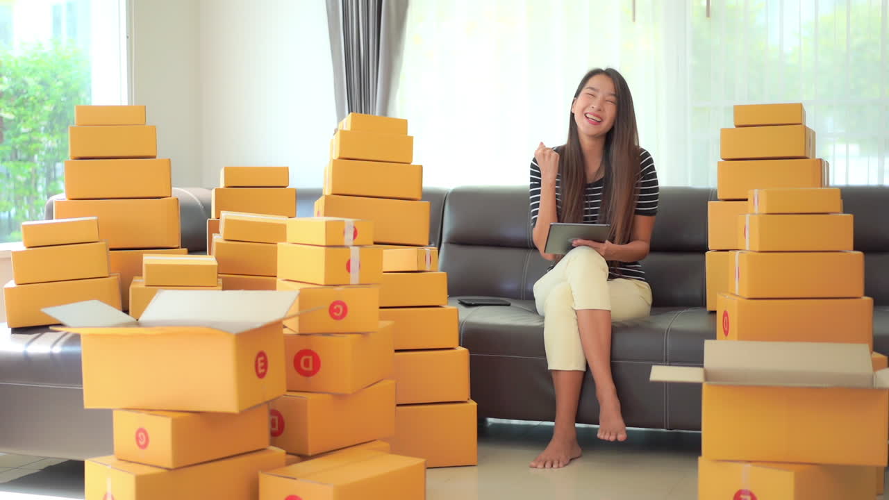 A young businesswoman sits on a couch surrounded by shipping boxes inputs data to her iPad, reacting to her successful input