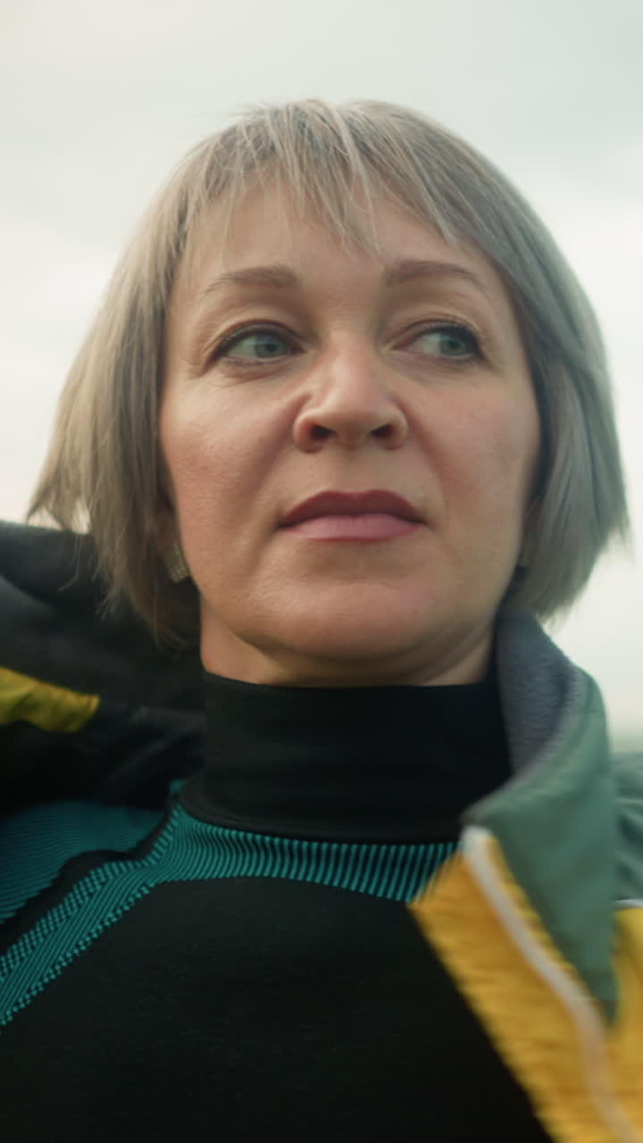primer plano de una mujer de mediana edad en traje verde y negro abriendo la chaqueta amarilla mientras mira a la distancia en un vasto campo abierto bajo un cielo nublado, con árboles verdes visibles en la distancia