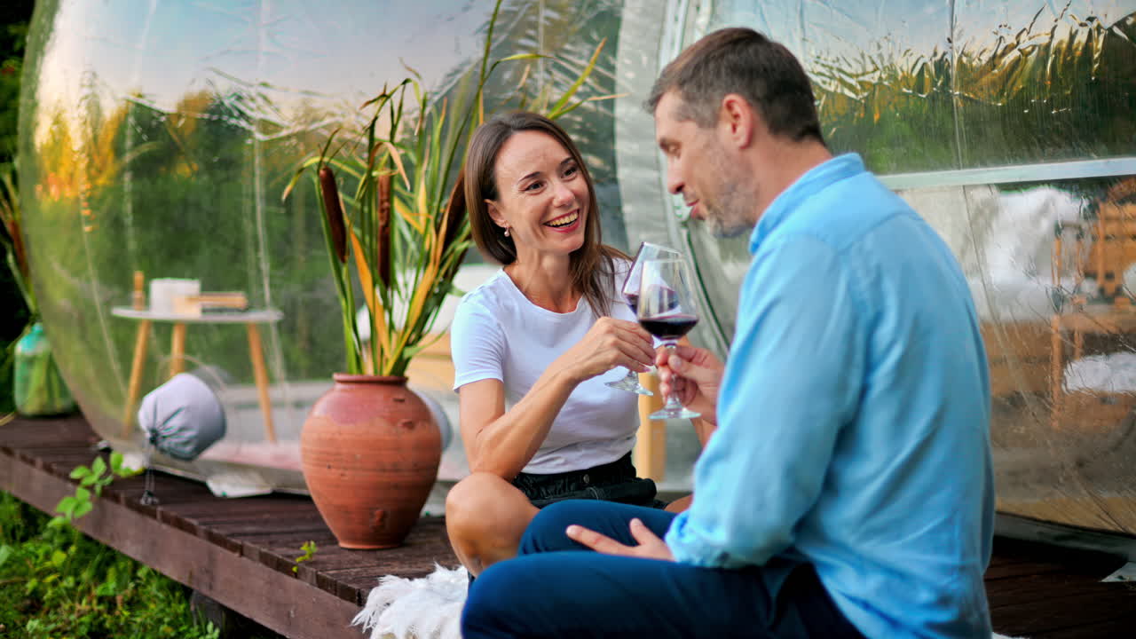 A happy couple drinking wine and talking near transparent bubble tent at glamping