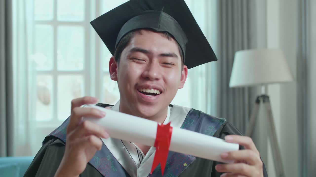 Excited Asian Man Showing Off A University Certificate During An Online Video Call. Male Graduate Wearing A Graduation Gown And Cap Sitting On The Living Room