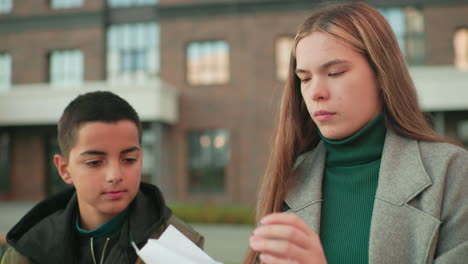 boy attentively watches as aunt demonstrates paper folding, nodding his head in understanding, showing moment of teaching, guidance, and learning outdoors with building background in natural light