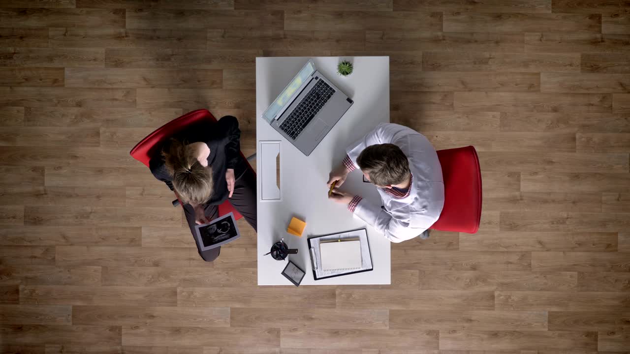 Læge udfører ultralyd på gravid ung mor, topshot, sidder ved bord med bærbar computer på kontoret