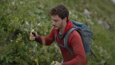 hiker in red jacket walking up a hill with hiking poles surrounded with green gass and alpine flowers